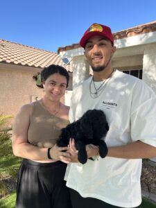 A couple smiling while holding a black puppy outside a house.