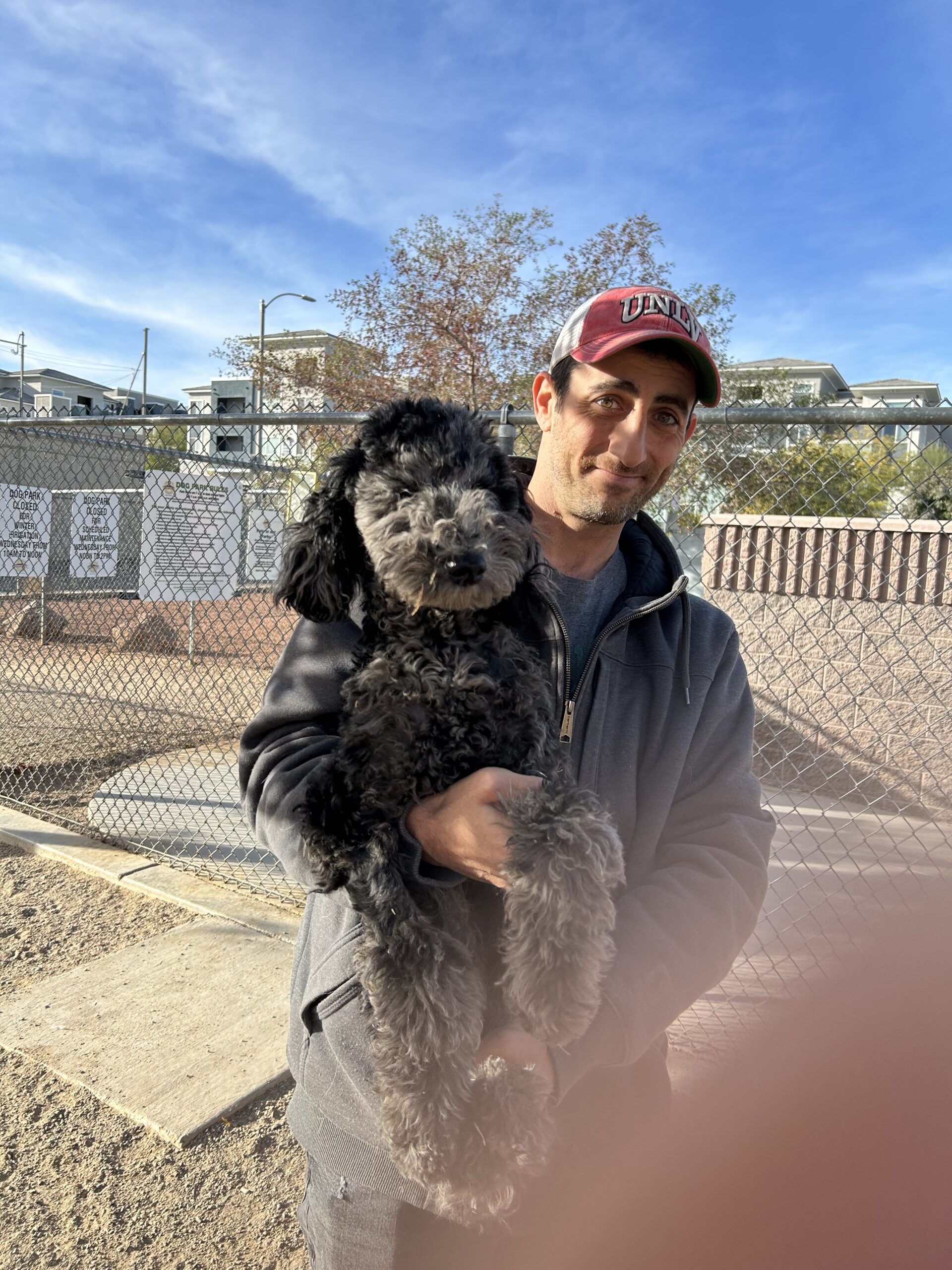 Man holding a black dog outdoors on a sunny day.